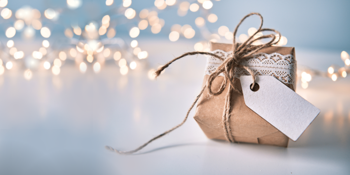 A gift box wrapped in tan paper with a twine bow and lace. There are twinkling lights in the background.