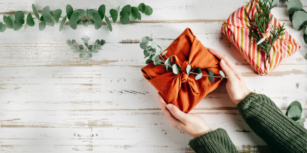 A woman holding a gift wrapped with fabric with eucalyptus on top. The color pallet is orange and green