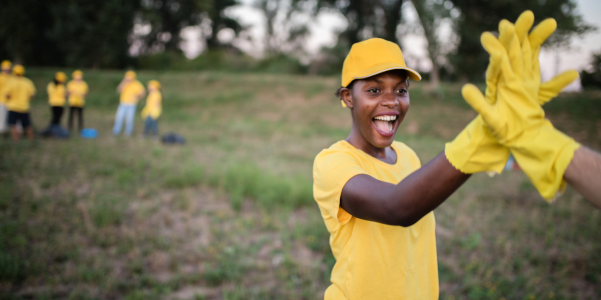 A young woman who is volunteering high-fives another volunteer who is behind the camera. They are wearing bright yellow shirts, hats, and gloves